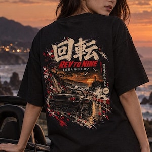 May include: A black short-sleeve t-shirt featuring a graphic print on the back. The design displays a black convertible car on a coastal road, Japanese kanji text, the words REV TO NINE, and a sunset scene with a lighthouse.