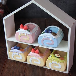 May include: Six miniature cake roll planters in a white wooden house-shaped shelf. The planters are decorated with frosting, sprinkles, and various toppings like strawberries, blueberries, and avocado.