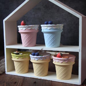 Könnte beinhalten: Ein weißes, hausförmiges Regal zeigt sechs eiscreme-förmige Pflanzgefäße. Die Pflanzgefäße sind in Pastellfarben gehalten und mit dekorativem Zuckerguss und Miniaturfrüchten verziert. Das Regal steht auf einer Holzoberfläche.