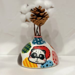 Puede incluir: Un jarrón de cerámica blanco con un diseño de cara de panda y patrones geométricos coloridos. El jarrón está decorado con lunares rojos, azules, amarillos, verdes y negros. El jarrón está lleno de bolas de algodón blancas y una piña marrón.