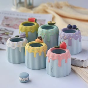 May include: Six pastel blue ceramic planters shaped like cupcakes with various toppings, including a strawberry, watermelon, avocado, blueberries, chocolate, and a macaron. The planters are decorated with colorful sprinkles.