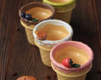May include: Four ceramic ice cream cone-shaped bowls with different toppings: blueberries, a cookie, an avocado, and a strawberry. A brown pretzel and coffee beans are in the foreground. The bowls have a yellow cone base and a white or pink rim.