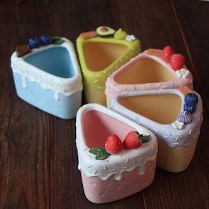 Pode incluir: Cinco vasos de cerâmica em forma de fatia de bolo de diferentes cores com diferentes coberturas, incluindo morangos, mirtilos, abacate e granulado. Os vasos estão dispostos sobre uma superfície de madeira.