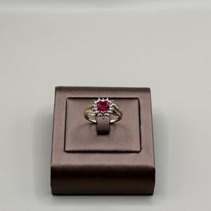 Könnte beinhalten: Ein goldener Ring mit einem herzförmigen roten Edelstein in der Mitte, umgeben von kleineren weißen Edelsteinen. Der Ring wird auf einer braunen Schmuckschatulle vor weißem Hintergrund präsentiert. Das Band des Rings hat ein filigranes Design.