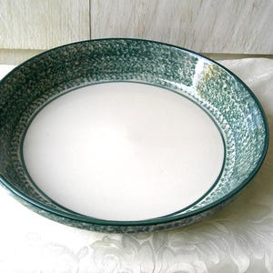 Può includere: Una ciotola rotonda in ceramica con interno bianco e bordo verde scuro strutturato. La ciotola è appoggiata su una tovaglia bianca con un sottile motivo floreale. La ciotola ha un diametro di circa 30 cm.