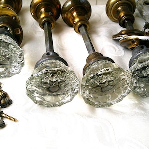 May include: Antique glass door knobs with brass hardware and a collection of brass screws. The knobs have a clear, faceted design. The hardware is tarnished, showing signs of age. A vintage hardware set.