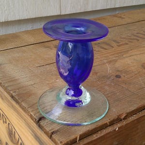 May include: A cobalt blue glass candle holder with a clear glass base. The candle holder has a flared top and a bulbous center. The base is a clear, flat circle. The candle holder is sitting on a wooden surface.