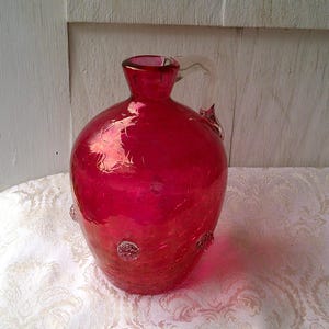 Puede incluir: Un jarrón de cristal rojo vibrante con un cuerpo redondeado y un cuello estrecho. El jarrón tiene un asa y adornos decorativos de cristal. El jarrón está sobre una tela blanca estampada.