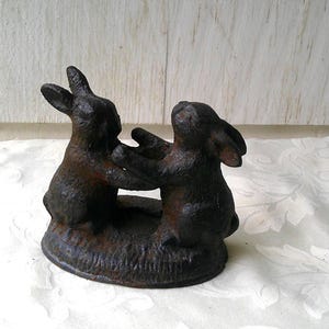 May include: A dark brown cast iron figurine featuring two rabbits standing on their hind legs and facing each other. The rabbits are positioned on a textured oval base, and the metal surface has a weathered, rustic appearance with visible texture.