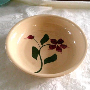 May include: A beige ceramic bowl with a floral design. The flower is burgundy with green leaves and stem. The bowl is round and has a smooth surface. The bowl is likely for serving food.