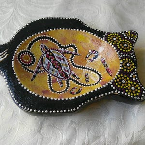 Puede incluir: Un plato decorativo con forma de pez con un diseño de arte aborigen. El plato presenta un borde negro con puntos blancos, un interior amarillo y naranja, y una tortuga y otros símbolos en el centro.