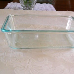May include: A rectangular, clear glass baking dish with a light blue rim. The dish is empty and sits on a white tablecloth with a floral pattern. The dish is approximately 23 cm long and 13 cm wide.