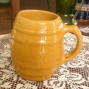 May include: A ceramic beer mug with a handle, in a warm yellow color. The mug has a barrel-like shape with horizontal ridges. It sits on a white lace tablecloth, suggesting a vintage or rustic theme.