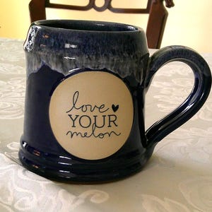 May include: A dark blue ceramic mug with a large handle. The mug has a cream-colored circle with the words "love YOUR melon" in black script. The mug is on a white tablecloth.