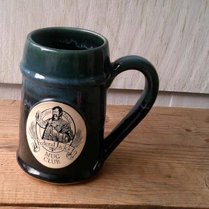 Puede incluir: Una jarra de cerámica de color verde oscuro con un asa grande. La jarra presenta un emblema circular de color crema con el texto "Federal Jack's Mug Club" y una ilustración de un hombre. La jarra está sobre una superficie de madera.