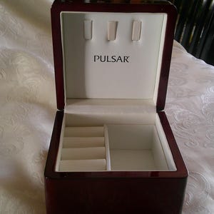 Może przedstawiać: Drewniana skrzynka w ciemnym kolorze czerwonym z białym wnętrzem i napisem "PULSAR" na pokrywie. Skrzynka ma przegrodę z czterema szczelinami i większą przegrodę.