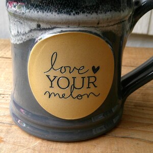 May include: A ceramic mug with a dark gray glaze and a large handle. A round, gold-colored sticker on the mug features the words "love YOUR melon" in black script. The mug sits on a wooden surface.