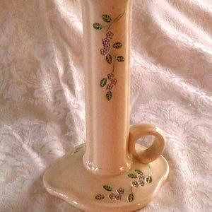 Puede incluir: Un portavelas de cerámica de color rosa pálido con un diseño floral. El portavelas tiene un solo asa y una base ancha.