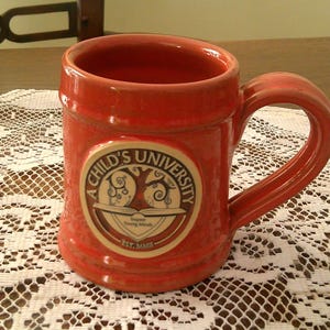 May include: A red ceramic mug with a large handle. The mug features a circular emblem with the text "A Child's University" and a tree design. The mug is sitting on a white lace tablecloth.
