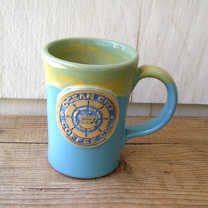 May include: A ceramic mug with a blue base, a green and yellow rim, and a curved handle. The mug features a circular emblem with the text "OCEAN CITY COFFEE CO." and a nautical design. The mug is on a wooden surface.