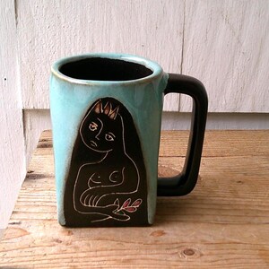 May include: A ceramic mug with a light blue glazed exterior and a black interior. The front features a dark, recessed panel with a white line drawing of a figure with a crown, holding a small plant with pink leaves. It has a black handle.