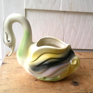 May include: A ceramic swan planter with a curved neck and open top. The swan is painted in shades of white, green, yellow, black, and pink. The planter is sitting on a wooden surface.