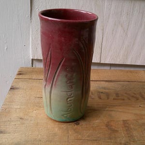May include: A handmade ceramic vase with a gradient of colors. The top is a deep burgundy, transitioning to a light green base. The word "Abundance" is inscribed on the side, with carved vertical lines. The vase is on a wooden surface.