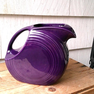 May include: A vibrant purple pitcher with a distinctive handle and spout. The ceramic pitcher has a glossy finish and a series of concentric ridges that encircle the body. The pitcher is set on a wooden surface.