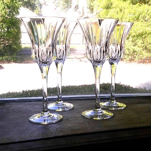 Può includere: Quattro flûte da champagne in cristallo trasparente con un design a coste. I bicchieri hanno un gambo alto e sottile e una coppa ampia e svasata. Sono disposti su una superficie di legno scuro davanti a una finestra con vista sul verde.