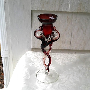 May include: A tall, elegant red glass candlestick holder with a twisted stem and a flared base. The top features a small, round bowl. The design includes decorative swirls and loops, creating a unique and artistic look. The candlestick holder is approximately 12 inches tall.