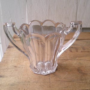 以下が含まれることがあります： 2つのハンドルが付いた、透明なヴィンテージガラスの砂糖入れ。ボウルはスカラップの縁とファセットベースを備えています。デザインは垂直線と底のスターバーストパターンが特徴です。木製の表面に置かれています。