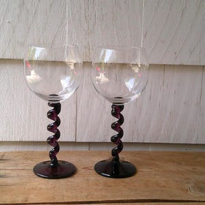 May include: Two clear glass wine glasses featuring deep purple stems with a distinct spiral, twisted design. Each glass has a wide, rounded bowl and a solid, circular purple base, providing a stable foundation for the elegant, elongated stem.