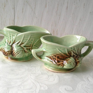May include: A ceramic creamer and sugar bowl set in a light green glaze with brown pinecone and needle relief detailing. The creamer features a curved handle and a pouring spout, while the sugar bowl has two side handles and a wide opening.