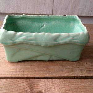 May include: A rectangular, light green ceramic planter with a textured, leaf-like design around the rim and sides. The planter is empty and sits on a wooden surface.