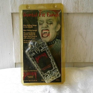Op de afbeelding: Een verpakking van "Dental Fit Fangs" met een doorzichtige plastic hoes. De verpakking toont een persoon met vampier make-up en de tekst "Best Selling Fangs in the World!" en "Classic Custom Fangs Scarecrow".