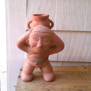 Puede incluir: Una escultura de terracota de una figura arrodillada con las manos en la cabeza, sosteniendo un pequeño jarrón. La figura tiene una cara simple y un cinturón. La escultura está sobre una superficie de madera.