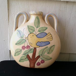 Könnte beinhalten: Eine cremefarbene Keramikkrug mit zwei Henkeln und einem bemalten Design. Das Design zeigt einen gelben Vogel mit blauen Flügeln, der auf einem braunen Ast mit grünen Blättern und roten Kirschen sitzt. Die Kanne hat einen runden Körper.