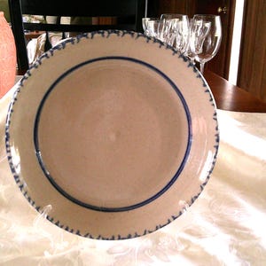 May include: A beige ceramic plate with a blue border and rim detail. The plate is on a white tablecloth. A pink vase and a Coca-Cola cup are also visible.