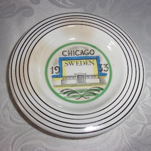 Puede incluir: Un platillo de cerámica blanca con círculos concéntricos negros alrededor del borde. En el centro hay una ilustración colorida de un edificio con el texto "CHICAGO SWEDEN 1933".