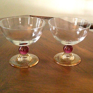 May include: Two clear glass champagne coupe glasses. Each glass has a round, purple stem and a circular, amber-colored base. The glasses are on a wooden surface.