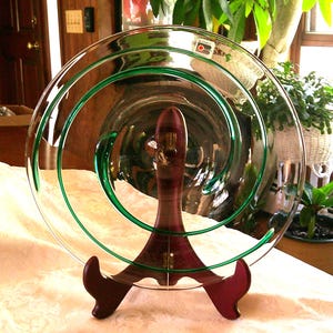 May include: A clear glass plate with a green spiral design on a wooden stand. The plate is round and has a diameter of approximately 12 inches.