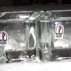 May include: Two clear glass candle holders with a square shape and a handle. Each holder has a Pilgrim Glass Handmade USA sticker. The glass has a slightly textured appearance.