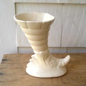 May include: Cream-colored ceramic cornucopia vase. The vase has a flared, scalloped rim and a spiraling, horn-like body. The base features decorative, stylized floral elements. The vase is displayed on a weathered wooden surface.