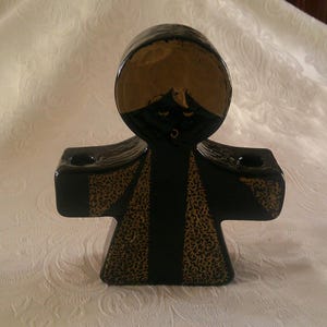 May include: Black and gold ceramic candle holder shaped like a person with a round head and two arms. The candle holder has two candle slots.