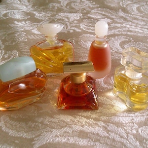May include: A selection of vintage perfume bottles in different shapes and sizes. The bottles contain amber-coloured liquid and have decorative stoppers. They are arranged on a patterned, off-white surface.