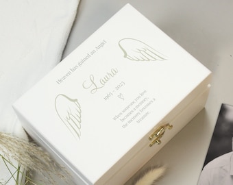 May include: White wooden keepsake box with a gold latch. The lid features a pair of angel wings and the text "Heaven has gained an Angel Laura 1965-2023 ♡ When someone you love becomes a memory, the memory becomes a treasure."