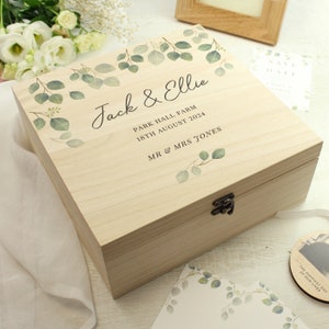 May include: A wooden keepsake box with a light wood finish and a hinged lid. The box is decorated with a eucalyptus leaf design and personalised with the text "Jack & Ellie", "Park Hall Farm", "18th August 2024", and "Mr & Mrs Jones".