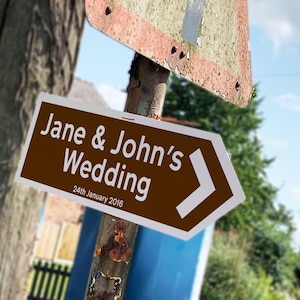 May include: A brown directional sign with white text that reads "Jane & John's Wedding" and "24th January 2016". The sign has a white arrow pointing to the right. The sign is mounted on a weathered metal pole.