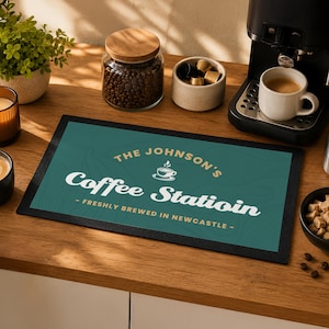 May include: A rectangular dark teal coffee station mat with a black border. The text reads THE JOHNSON'S Coffee Statioin with a small coffee cup icon and the text FRESHLY BREWED IN NEWCASTLE. The mat features a subtle wavy line pattern.