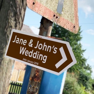 Puede incluir: Un letrero direccional marrón con texto blanco que dice "Jane & John's Wedding" y "24th January 2016". Una flecha blanca indica la dirección. El letrero está montado en un poste de metal desgastado.